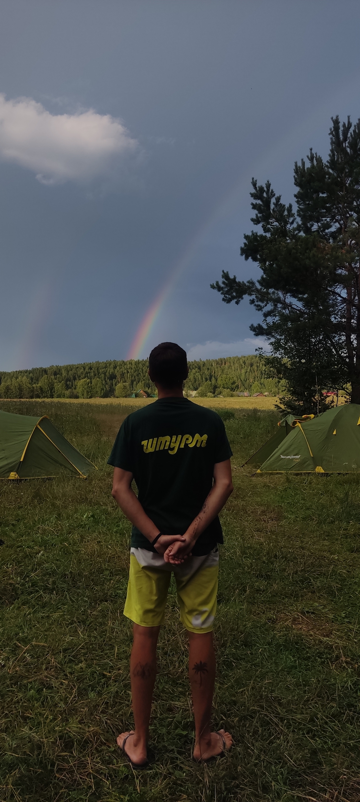 Турист в лагере на берегу реки наблюдает радугу после дождя рядом с палатками